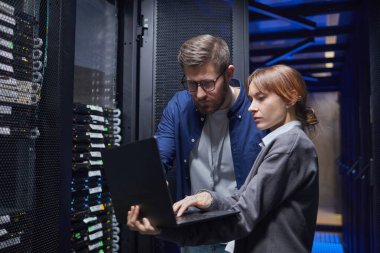 Bir veri merkezindeki sunucu bağlantılarını inceleyen iki bilişim uzmanı, sorun çıkarmak için gerekli adımları tartışırken dizüstü bilgisayara odaklanıyor