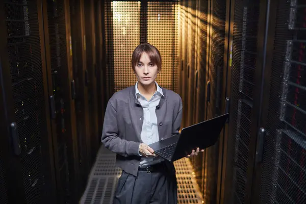Bilgi merkezinde çalışan beyaz bir kadının portresi sunucu rafları arasında dizüstü bilgisayarını tutarken arka planda görünür.