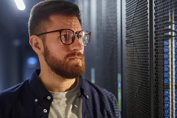 Bir veri merkezinde duran sakallı adam, sunucular ve ekipmanlarla çalışıyor, gözlük takıyor ve odaklanmış görünüyor. Teknoloji ve sunucular arka planda görünür