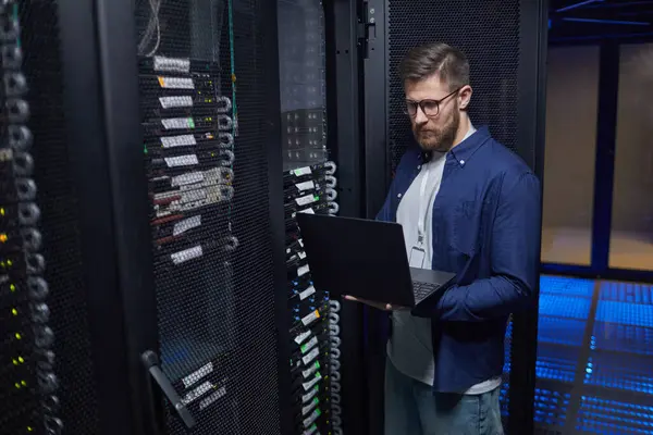 Modern sunucu odasında elinde dizüstü bilgisayarla duran erkek bilişim teknisyeni ağ ekipmanlarını kontrol ediyor ve düzgün bir işlevsellik sağlıyor. Sunucu rafları ve veri kablolarının yer aldığı yüksek teknoloji ortamı