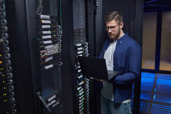Мужчина IT-техник, стоящий в современной серверной комнате с ноутбуком, проверяющий сетевое оборудование и обеспечивающий надлежащую функциональность. Высокотехнологичная среда с серверными стойками и кабелями передачи данных