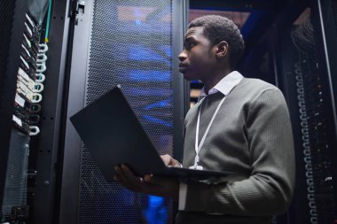 Afro-Amerikalı adam sunucu odasında elinde dizüstü bilgisayar ve izleme cihazlarıyla dikiliyor. Ağ oluşturma araçlarına odaklanarak düzgün işletim ve sistem performansı sağlamaya çalışıyor.