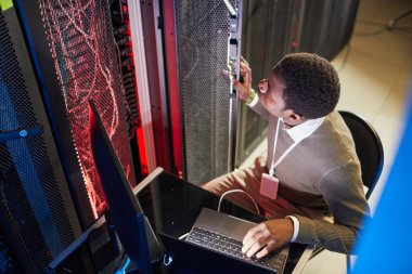 Veri merkezindeki sunucu odası ekipmanlarını izleyen Afro-Amerikalı bir adam dizüstü bilgisayarla oturup sistemlerin çalışmasını ve güvenliğini sağlıyor.