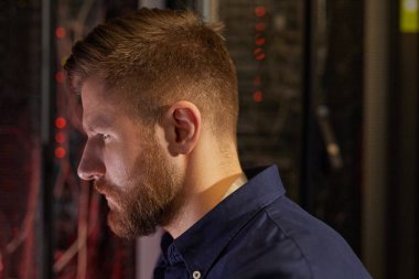 Sakallı bir adam veri merkezindeki sunucuları dikkatle inceliyor mavi gömlekli, yan profil görünümlü ve iş sırasında yakalanan davranışları olan.