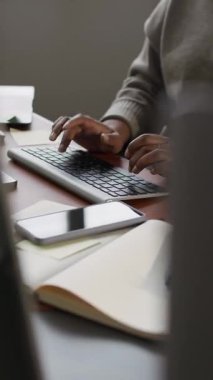Ofis masasında çalışan tanınmayan bir ofis çalışanının ellerinin dikey kesilmiş görüntüsü. Yakınlarda açık bir telif defteri ve cep telefonu var.