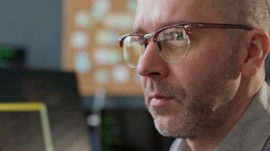 Gözlüklü, modern ofis masasında bilgisayar önünde oturan ve program kodlaması üzerinde çalışan konsantre orta yaşlı beyaz erkek geliştiricinin yakın plan fotoğrafı.