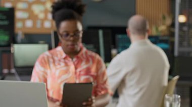 İyi donanımlı açık uzay ofisinde dijital tablet üzerinde çalışan genç siyahi kadın programcının portresini yakınlaştırın ve arka planda erkek meslektaşı kodlayan kameraya bakın.