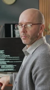 Gündelik giyimli, kendinden emin orta yaşlı beyaz erkek programcının dikey portresi sandalyeye dönüyor ve modern ofiste bilgisayarların önünde program kodlarıyla poz veriyor.