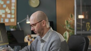 Modern ofis masasında çalışan, odaklanmış orta yaşlı, gözlüklü beyaz erkek geliştiricinin yavaş yavaş portresi, sonra bulanık arka planda program kodlarıyla çalışan kameraya bakıyor.