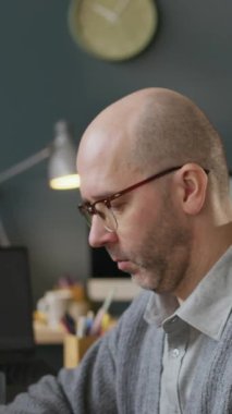 Ofis masasında çalışan, konsantre olmuş, orta yaşlı, gözlüklü beyaz erkek geliştiricinin dikey yavaş portresi, sonra da bulanık arka planda program kodlarıyla çalışan kameraya bakıyor.