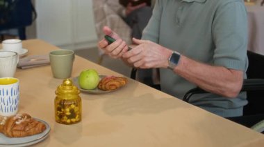 Çay, meyve ve hamur işleriyle dolu masada akıllı telefonuna odaklanan beyaz gözlüklü üst düzey bir adam, arka planda kitap okurken, her ikisi de rahat huzurevinde yaşıyor.