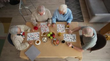 Öğleden sonra huzur evinin güneşli yaşam alanında çay, meyve ve hamur işleriyle dolu masada tombala oynayan dört son sınıf öğrencisinin tepeden tırnağa görüntüsü.