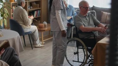 Genç siyahi kadın bakıcı tekerlekli sandalyedeki yaşlı beyaz adamın akıllı telefon tutmasına yardım ederken diğer sakinler rahat, güneşli bir huzurevinde dinleniyorlar.