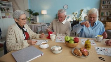 Üç yaşlı arkadaş, çay, hamur işi ve meyve dolu masada tombala oynuyorlar rahat, bitkilerle dolu yaşlılar için yaşam alanı.