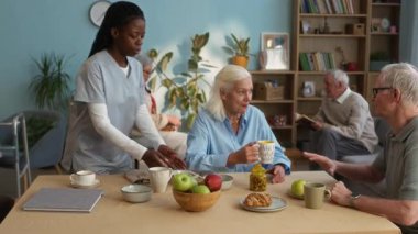 Genç siyahi kadın bakıcı bulaşık yıkarken yaşlılar sıcak içecekler, meyve ve hamur işleriyle dolu, rahat bir huzurevinde sabahın tadını çıkarırken.
