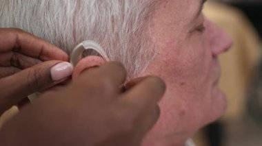 Bakıcının tanınmayan elleri, yaşlı bir adamın kulağına nazikçe işitme cihazını yerleştiriyor.
