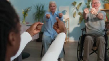 Genç siyahi kadın bakıcı tarafından yönetilen grup egzersizine katılan huzurevi sakinleri, önlerinde oturuyor ve el ve kol egzersizi yapıyorlar.