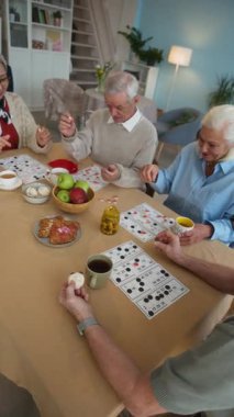 Dört neşeli son sınıf öğrencisinin tombala oynamaktan ve içtenlikle sohbetten keyif aldıklarının dikey çekimi, çay saatinde, huzur evi ortamının ışıl ışıl, ferah oturma odasında otururken