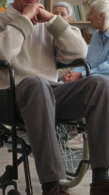 Tekerlekli sandalyede oturan Kafkasyalı yaşlı bir adamın dikey portresi, sakinleri sıcak, huzur evi ortamında sohbet ederken sakince oturuyor.