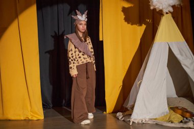 El yapımı kuş tüyü kostüm giyen küçük kız çeşitli yetenekleri olan çocuklar için tiyatroda wigwam ile süslenmiş sahnede dururken Amerikan yerlilerinin kültürü etrafında oyun oynuyor.