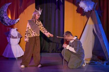 Çocuk tiyatrosunun seyirci alanından uzun bir çekim. Yenilen şövalye dizlerinin üzerine çöküp sahne ışıkları altında kadın savaş galibi önünde duruyor.