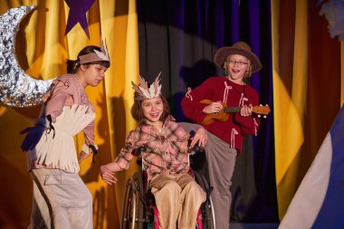 Tekerlekli sandalye kullanan neşeli genç bayan aktör, ukulele 'ye şarkı söyleyen arkadaşlarıyla birlikte çocuk tiyatro okulunda Batı müzikali oyununa katılıyor.