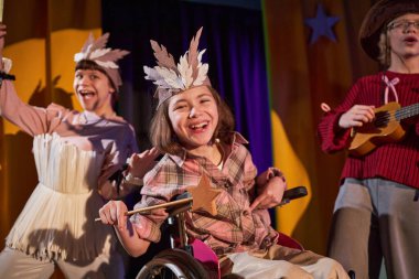 Çocuk tiyatrosunda büyücüyü oynayan tekerlekli sandalyeli genç bir kızın portresi elinde tahta sihirli sopayla sahne ışıkları altında bir grup arkadaşla şarkı söylerken