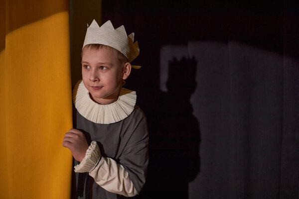 Medium shot of cute little boy wearing handmade paper crown looking from behind yellow curtains with anticipation while acting on stage of childrens theater, copy space