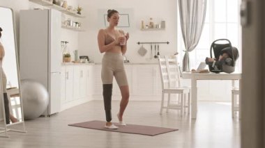Modern bacak protezi olan zayıf, sportif bir kadının evde yoga minderinin üzerinde durup su içerken ve bebek koltuğunda yatarken bebeğini kontrol ederken geniş açılı görüntüsü.