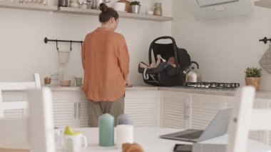 Orta büyüklükte, beyaz kadın, mutfak atölyesinde kahvaltı hazırlarken güvenli bebek koltuğunda yatarken ve dizüstü bilgisayarda klavyeyle yazarken.