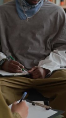 Modern sınıfta, grup terapisi sırasında sandalyede oturan ve diğer katılımcıları gözlemlerken düşüncelerini panoya yazan Afrikalı Amerikalı gencin dikey yatay pozu.