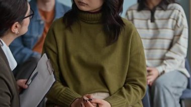 Sınıfta grup terapisi sırasında arka planda konuşan farklı akranları olan üzgün Asyalı genç kızı destekleyen empatik kadın psikoloğun odak noktası.