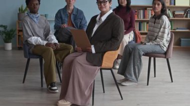 Çok ırklı dört gencin grup portresi ve kendine güvenen melez bir kadın psikolog fotoğraf makinesine poz veriyor ve rahat modern sınıflarda daireler çiziyor.