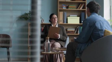 Genç melez kadın psikoloğun dosyalama tahtası tutarak rahat bir ofiste bireysel danışmanlık sırasında önünde oturan genç adamla konuşmasının yavaş çekimleri.