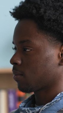 Kendine güvenen Afro-Amerikan ergen çocuğun dikey yakın çekimi modern sınıfta oturuyor ve elinde pano tutarak yürüyen ve tanınmayan öğretmene bakıyor.