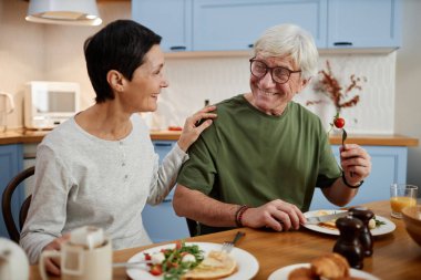 Yaşlı bir çift, rahat bir mutfakta yemek yiyor, keyiflerini paylaşıyorlar. Yeşil gömlekli adam çatalla yemek tutuyor, kadın gülümsüyor ve omzuna dokunuyor.