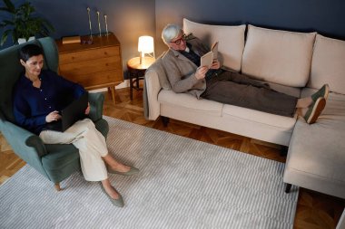 Son sınıflar modern dekorlarla çevrili dijital cihazlar ve kitaplarla oturma odasının keyfini çıkarıyorlar. Bir kişi dizüstü bilgisayarı incelerken diğer kişi okuyor.