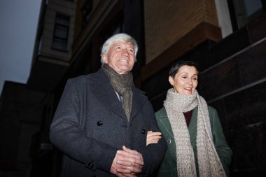 Işıl ışıl kentsel ortamda yürürken gülümseyen yaşlı bir çift, kalın ceketler ve eşarplar giyiyorlar. Gece ayarı çiftler arasında paylaşılan ana derinlik eklemesi