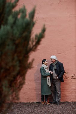 Yaşlı çift, uzun yeşillik ve duvarın yakınındaki çakıl yolda bardakları tutarken sohbet ediyorlar, kışlık giysiler giyiyorlar, mutlu ve mutlu görünüyorlar.