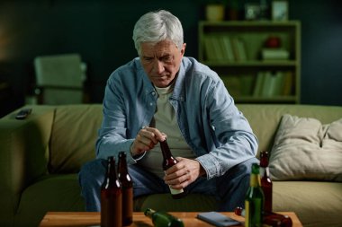 Kanepede oturan yaşlı bir adam elinde bira şişesiyle oturma odasındaki diğer şişelerle birlikte düşünceli bir ifadeyle oturuyor.