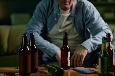 Birkaç alkollü içecekle masada oturan orta yaşlı bir adam düşünceli görünüyor ve loş ışıklandırılmış oda arka planına yansıyor.