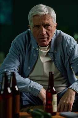 Beyaz saçlı yaşlı bir adamın portresi bir barda oturuyor, elinde bir şişe, ön planda bira şişeleriyle, doğrudan kameraya bakıyor.