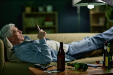 Rahat bir oturma odasında kanepede uzanmış akıllı telefonuna göz gezdiriyordu. Öndeki masada soda şişeleri, uzaktan kumanda ve kitaplar var.