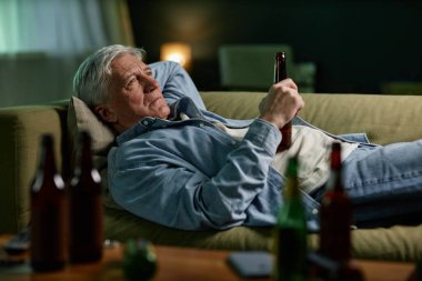 Yaşlı bir adam loş bir odada içki içerken kanepede uzanıyor, bir rahatlama ve içgözlem anı yakalıyor.