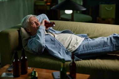 Yaşlı adam boş şişelerle çevrili elinde içki tutarken kanepede uzanıyor. Gündelik ev ortamında akşam rahatlığının keyfini çıkarıyor gibi görünüyor.