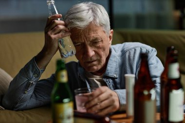 Gri saçlı, elinde boş bir şişeyle sorunlu görünen yaşlı bir adam. Çeşitli alkol şişeleriyle çevrili, derin düşüncelere dalmış ve kaybolmuş görünüyor.