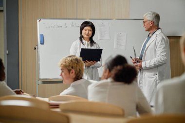 Asyalı bayan tıp öğrencisinin portresi beyaz tahtanın yanında duruyor ve konuyu sunarken kıdemli profesör gözlemliyor, boşluğu kopyala