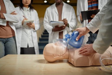 Tıp fakültesinde ilk yardım eğitimi sırasında çanta kapakçığı maskeli suni teneffüs mankeninin aşırı yakın çekimi, not alan öğrencilerin bulanık geçmişi