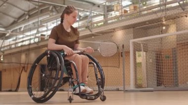 Alçak açılı, tekerlekli sandalyedeki beyaz kız çocuğu spor salonunda antrenmandan önce badminton raketiyle ısınıyor.