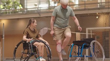 Tekerlekli sandalyede oturan, engelli genç bir kızın yanında oturan, kapalı sahada raketle badminton öğreten kıdemli Kafkas koçu.